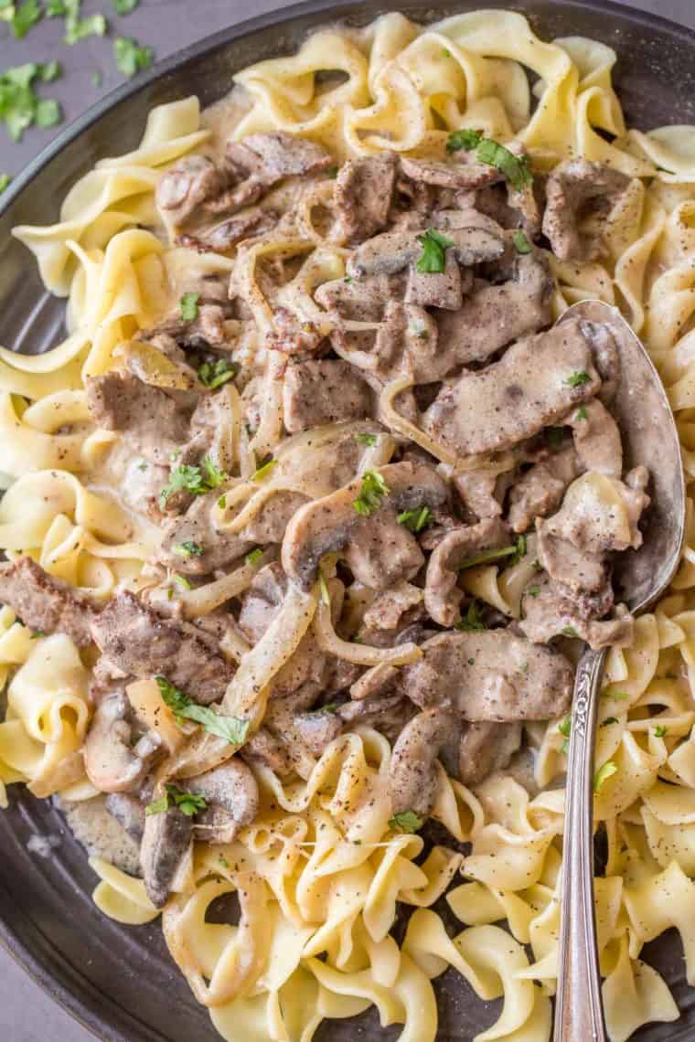 Beef stroganoff on top of egg noodles on a black plate with a spoon topped with fresh greens. 
