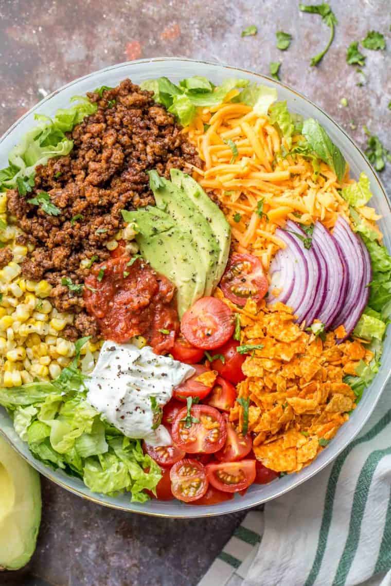 Dorito taco salad in a bowl unmixed topped with fresh greens next to an avocado.