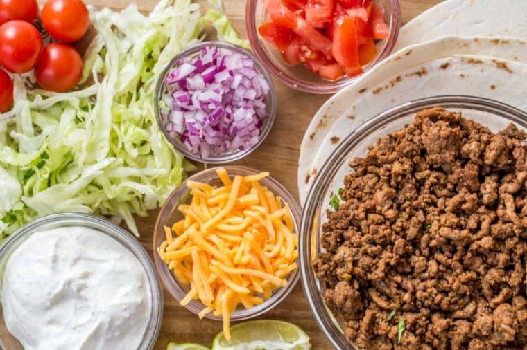 Ground taco meat, sour cream, cheese, onions, tomatoes, shredded cheese all in small glass bowls laid out on a platter. 