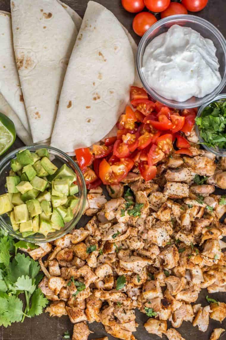 Seasoned chopped chicken, tomatoes, avocado, tortillas, sour cream all laid out on a platter. 