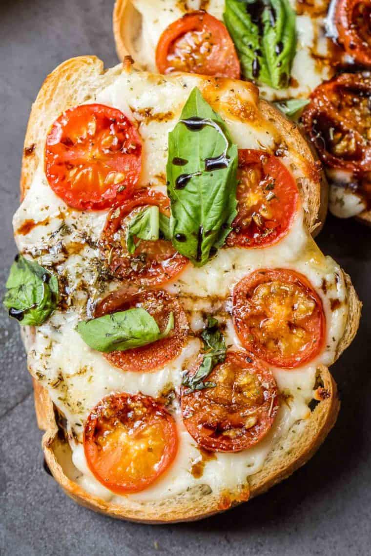 A slice of bread topped with melted cheese, sliced tomatoes, chopped basil, and drizzled with balsamic glaze.
