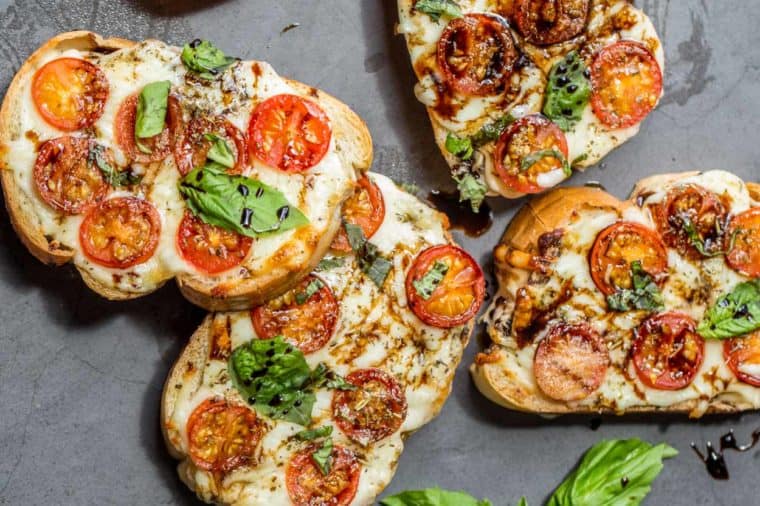 Four Margherita pizza toast laid out on a board drizzled with balsamic glaze and fresh chopped basil. 