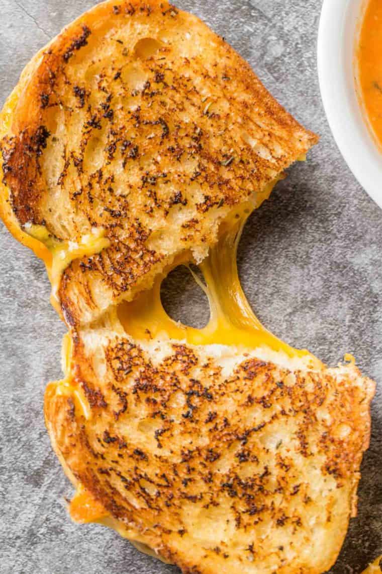 Grilled cheese cut in half with cheese oozing from the middle next to a bowl of tomato soup.