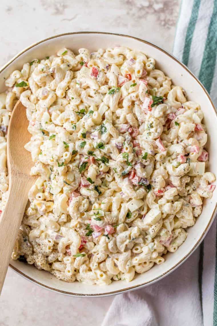 Macaroni pasta salad in a white bowl with a wooden spoon topped with fresh chopped greens.