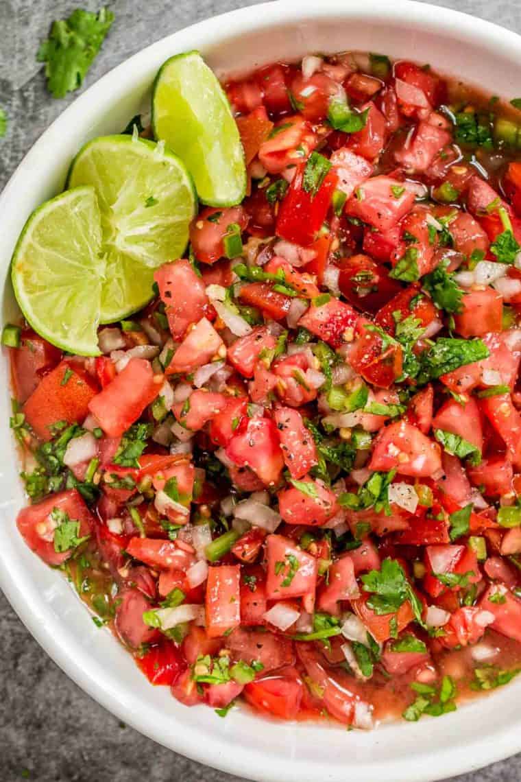 Homemade pico de gallo in a white bowl with lime slices topped with fresh chopped greens.