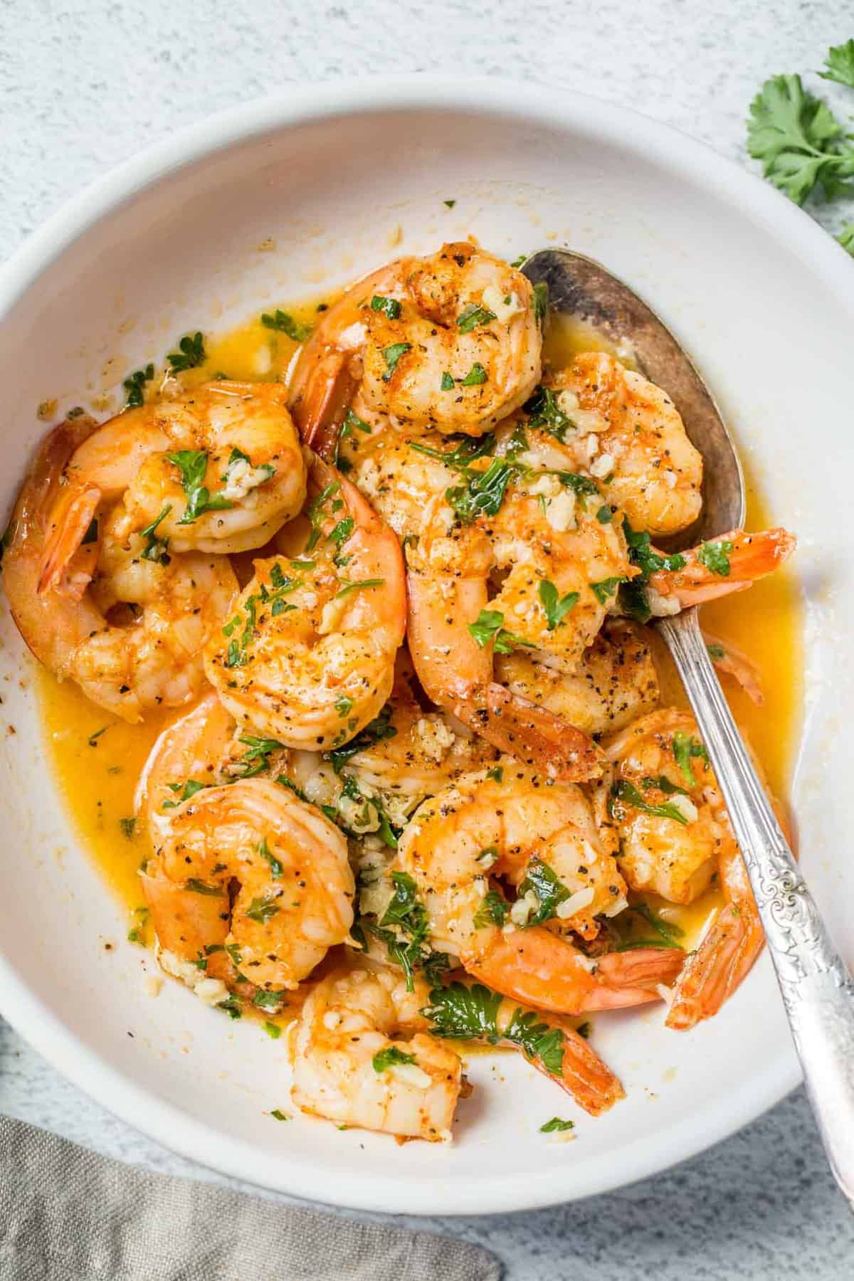 A white bowl loaded with shrimp scampi with a metal spoon topped with fresh chopped parsley.