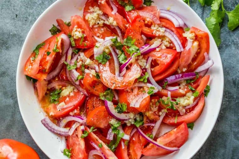 Fresh tomato salad in a white bowl loaded with herbs and red onion next to cilantro and a tomato.