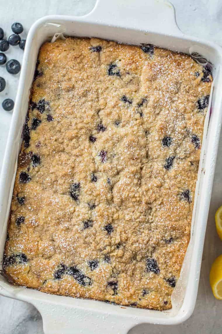 Blueberry cake in a white casserole dish topped with powdered sugar next to fresh blueberries and lemons.