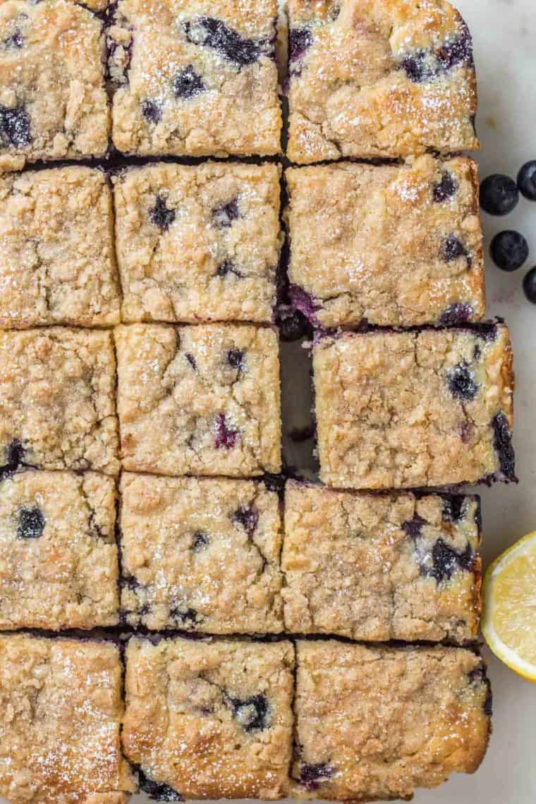 Blueberry cake sliced into squares on a platter topped with powdered sugar next to a lemon.