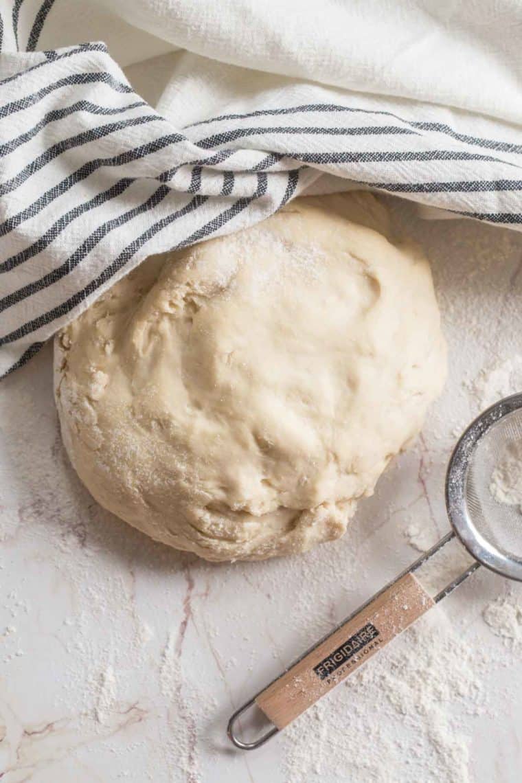 Pierogi dough sprinkled with flour next to a sieve and a striped white rag. 