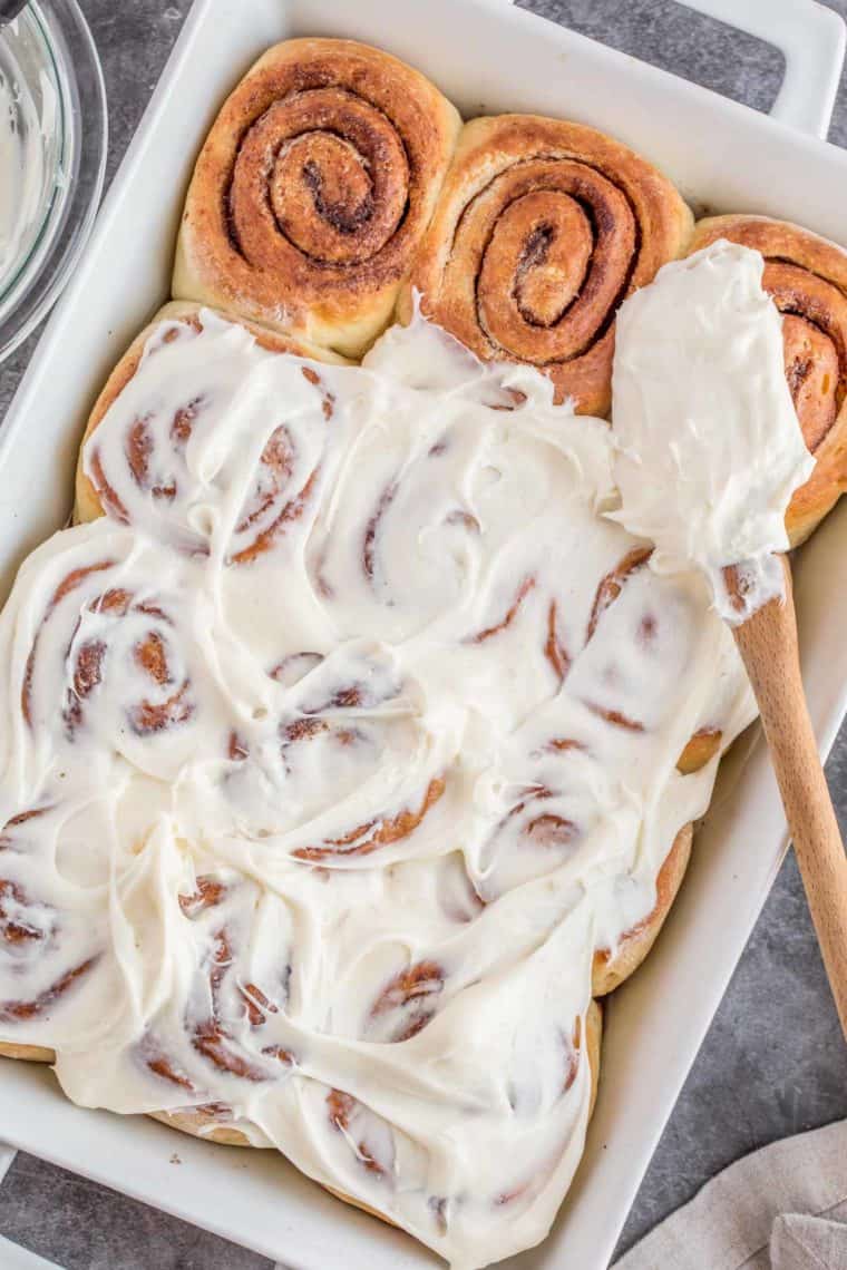 White casserole dish loaded with cinnamon rolls topped with cream cheese frosting and a spatula.