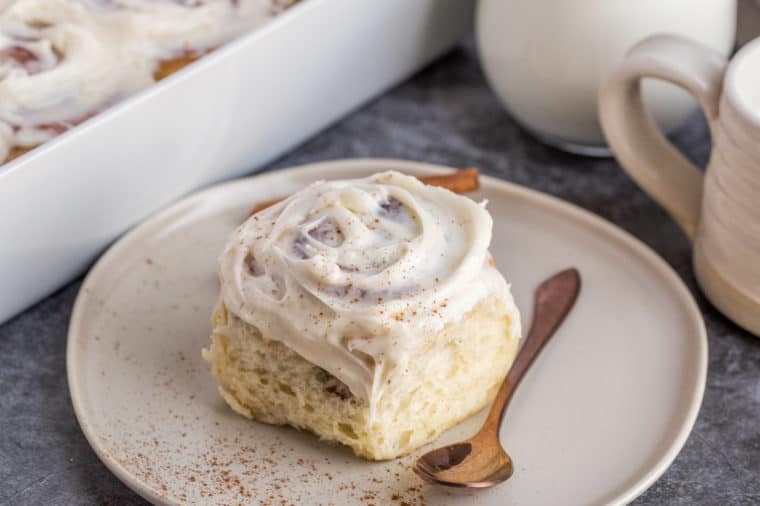 A cinnamon roll on a plate topped with cinnamon with a spoon next to a casserole dish and cups.