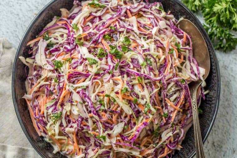 A black bowl loaded with coleslaw and topped with fresh chopped greens and a metal spoon.