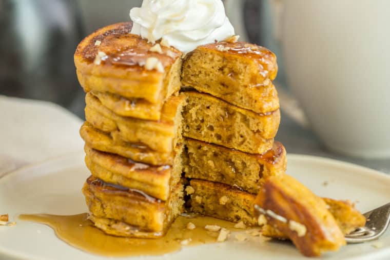 A stack of pumpkin pancakes on a plate topped with whipped cream, syrup and walnuts with a fork. 