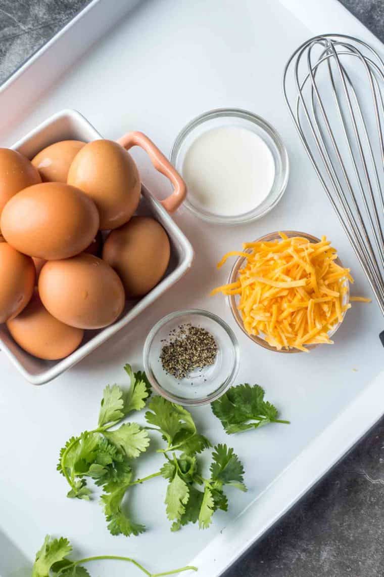 A white baking sheet with a carton of eggs, cilantro, cheese, seasonings and a whisk.