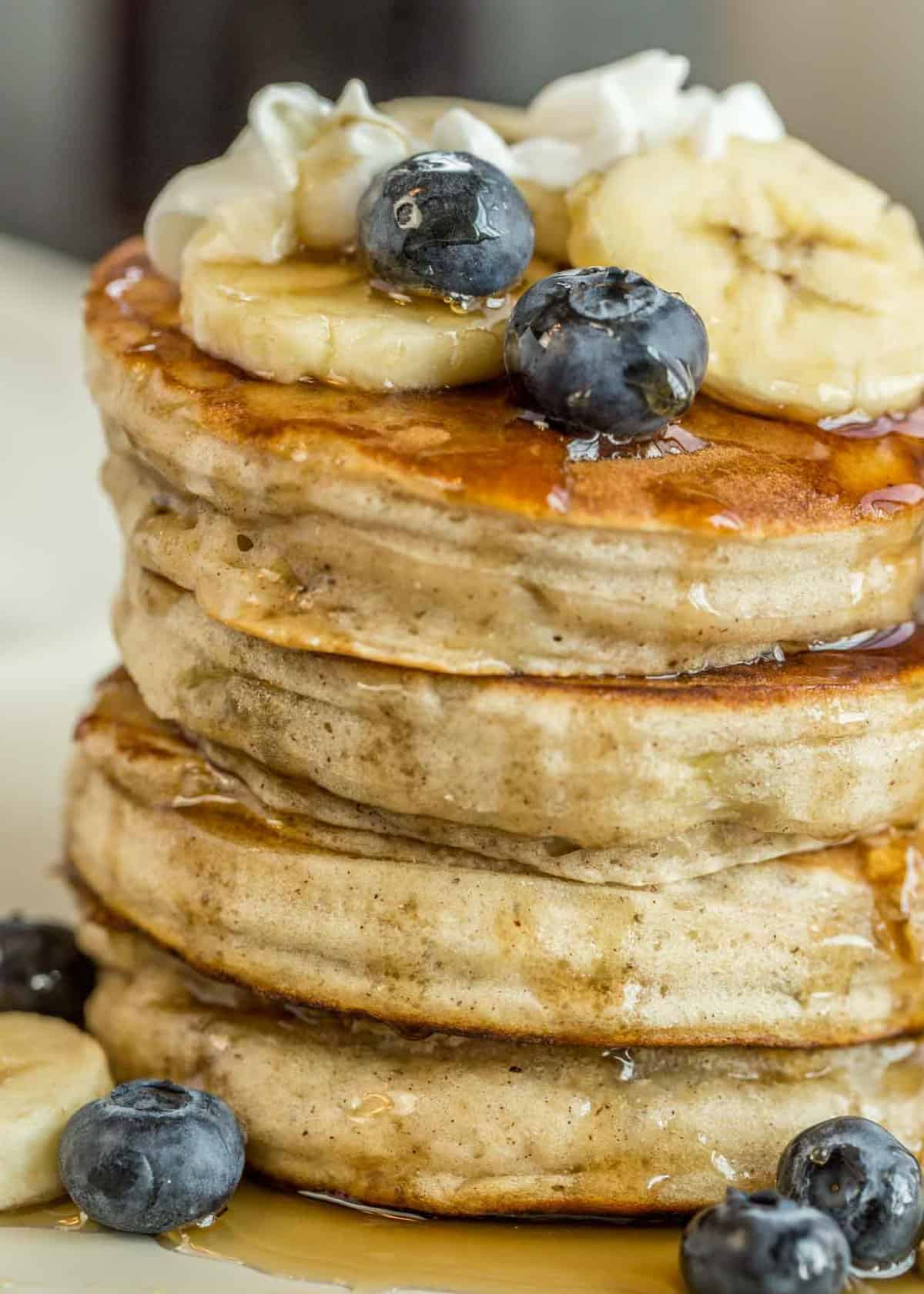 Banana pancakes stacked on top of each other with fresh bananas, blueberries, whipped cream and syrup.