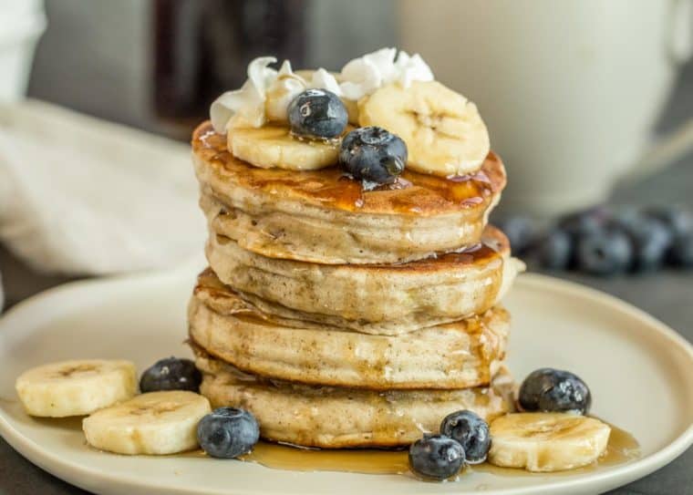 Banana pancakes on a plate with fresh blueberries, bananas, whipped cream and syrup.