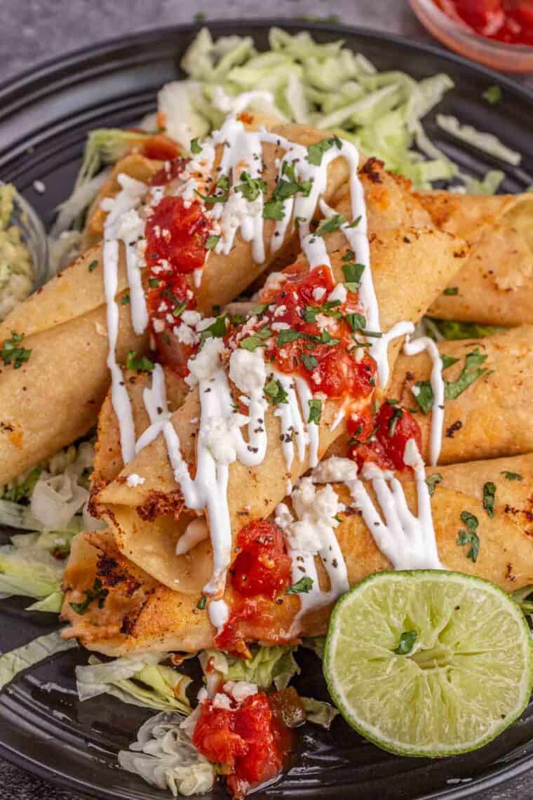 Up close picture of taquitos topped with sour cream, tomatoes, and fresh chopped greens. 