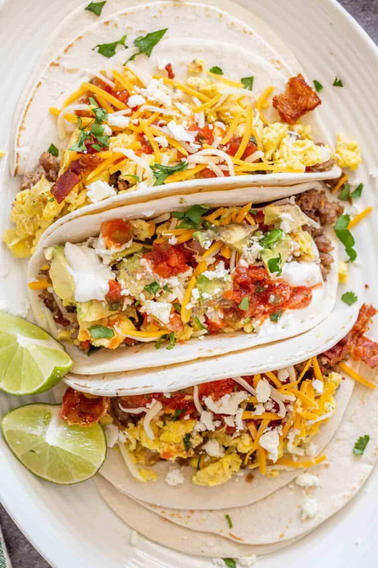 Three breakfast tacos on a white plate loaded with toppings and topped with greens.
