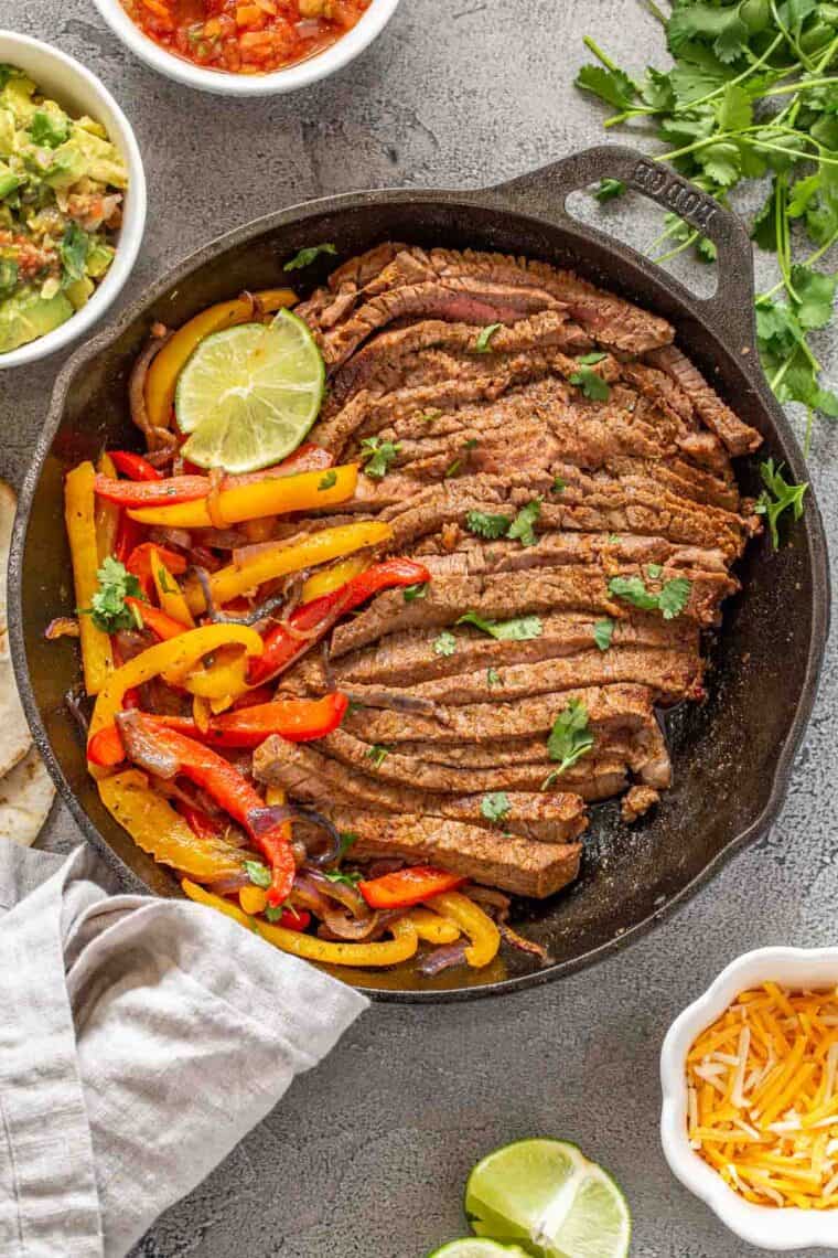 Beef steak fajitas in a black cast iron with veggies next to the toppings.