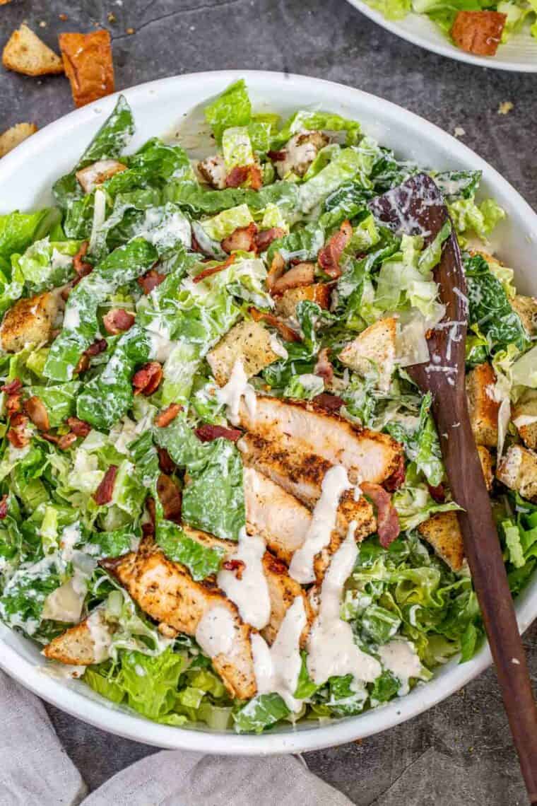 Caesar salad in a white bowl with a wooden spoon loaded with bacon and chicken. 