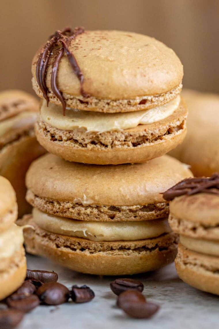 Coffee macarons stacked on top of each other next to other macarons and coffee beans. 