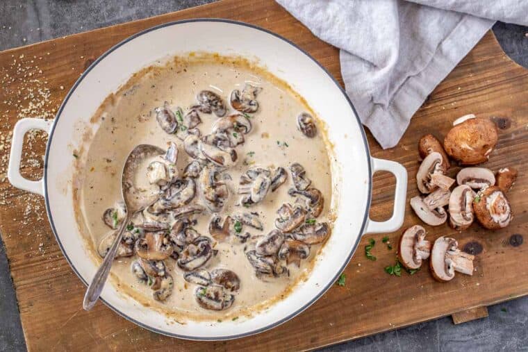 Mushroom sauce in a white skillet on a cutting board with mushrooms and a white rag. 