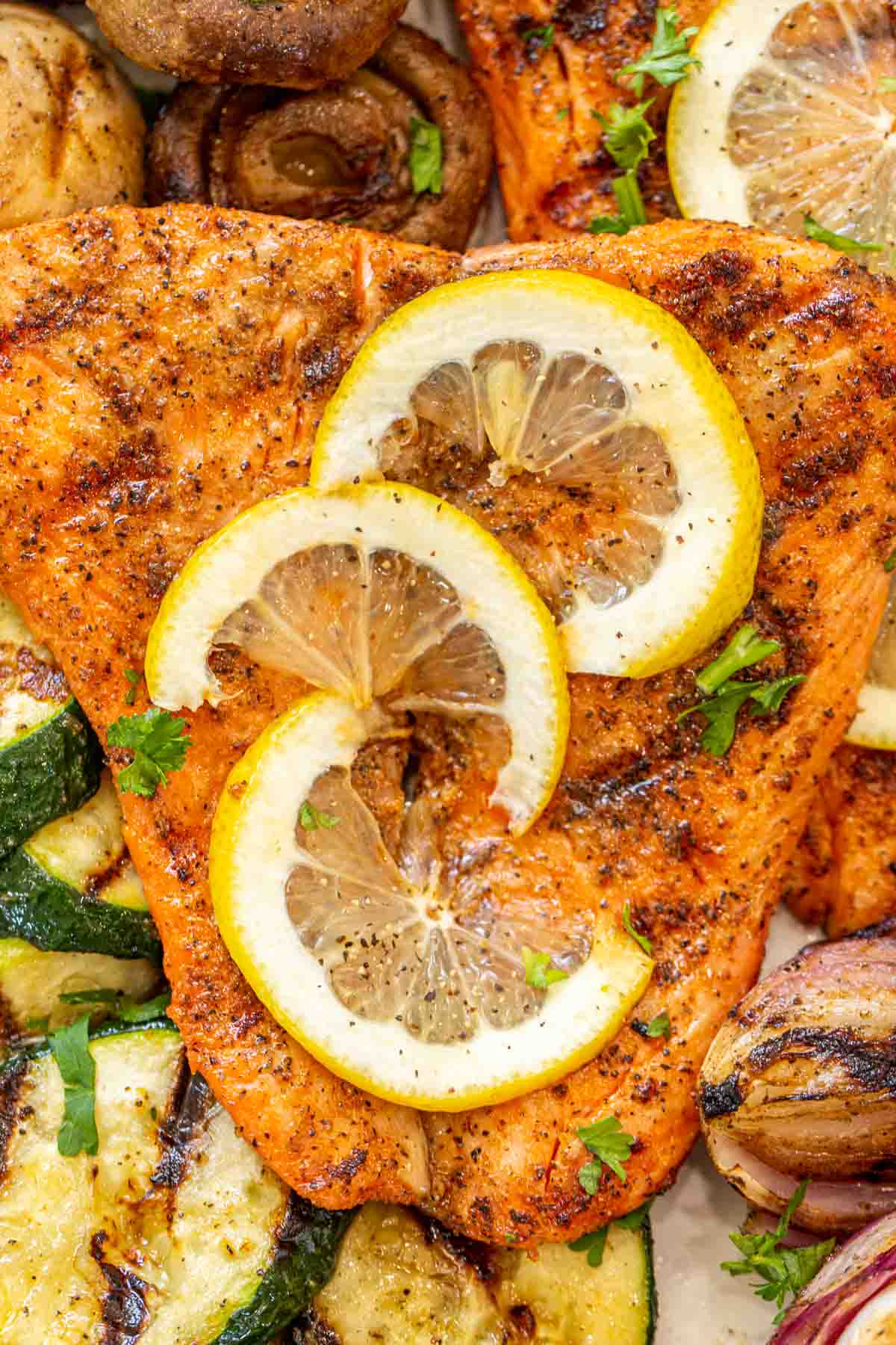 Salmon fillets topped with lemon slices and fresh chopped greens next to grilled vegetables. 