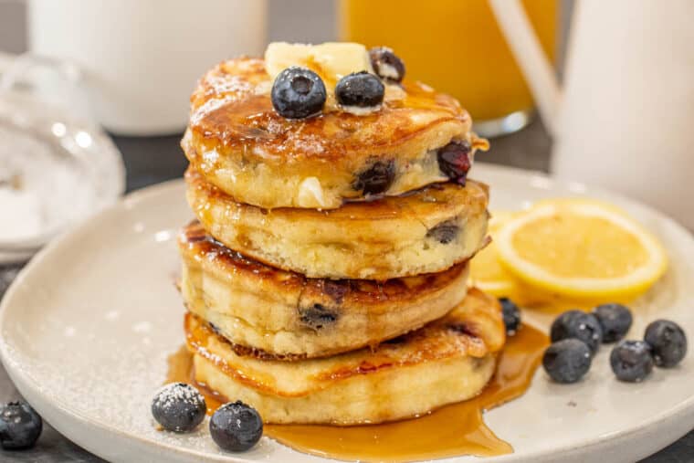 A stack of lemon blueberry pancakes, decorated with fresh blueberries and drizzled with maple syrup.