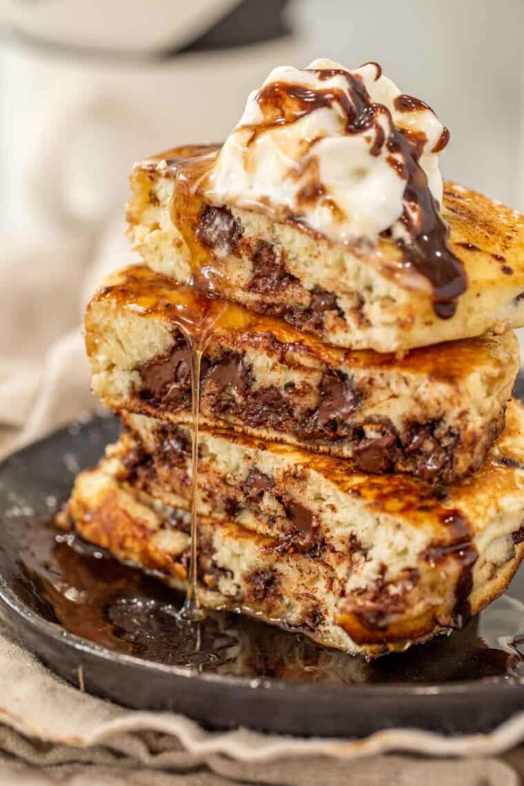 Chocolate chip pancakes cut in half stack on a plate with whipped cream and drizzled syrup. 