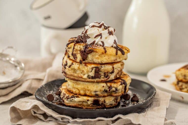 Chocolate chip pancakes stacked on a black plate topped with whipped cream.