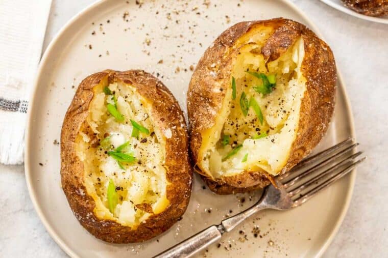 Two baked potatoes filled with butter and green onions on a plate with a fork.