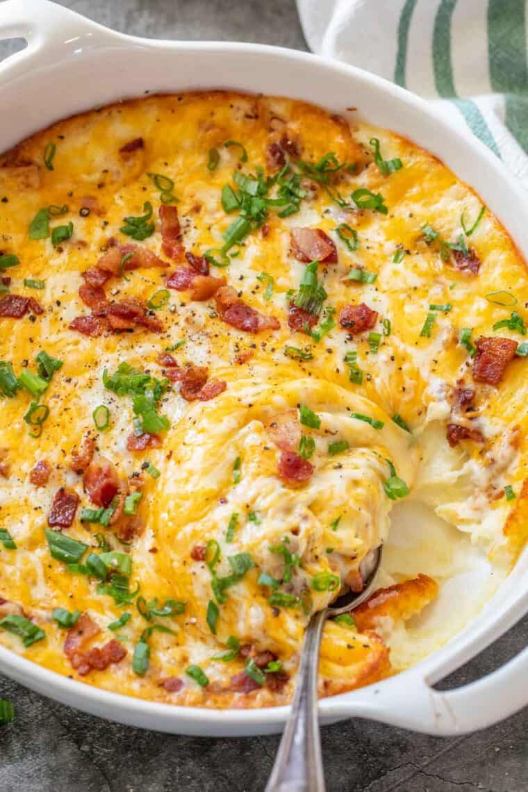 Loaded baked potato casserole in a white casserole dish with a spoon inside and topped with bacon and green onion.