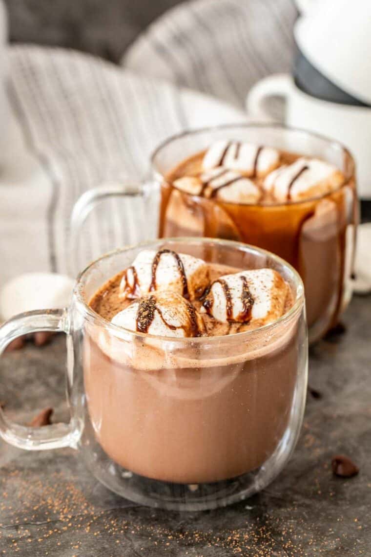 Two glass mugs of hot chocolate topped with large marshmallows and chocolate drizzle. 