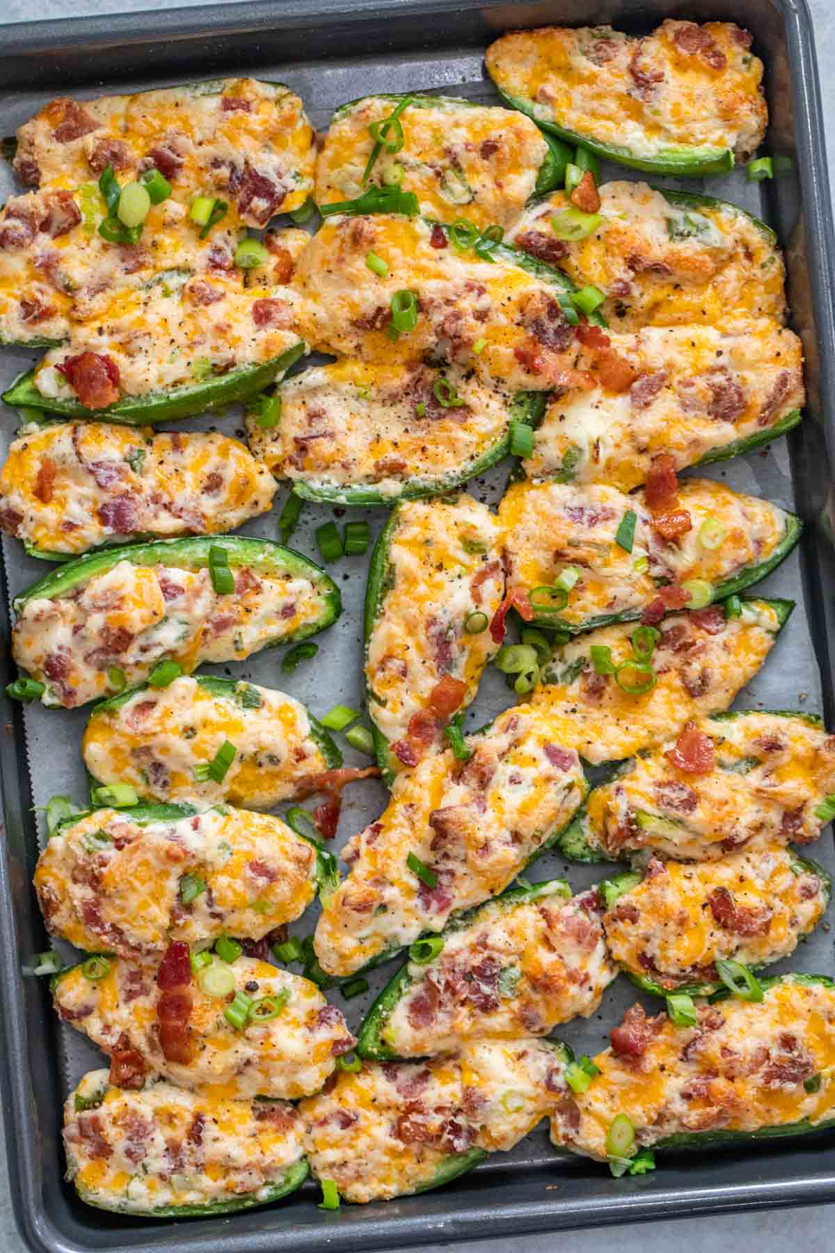 Baked jalapeno poppers topped with green onion and bacon laid out on a baking sheet. 