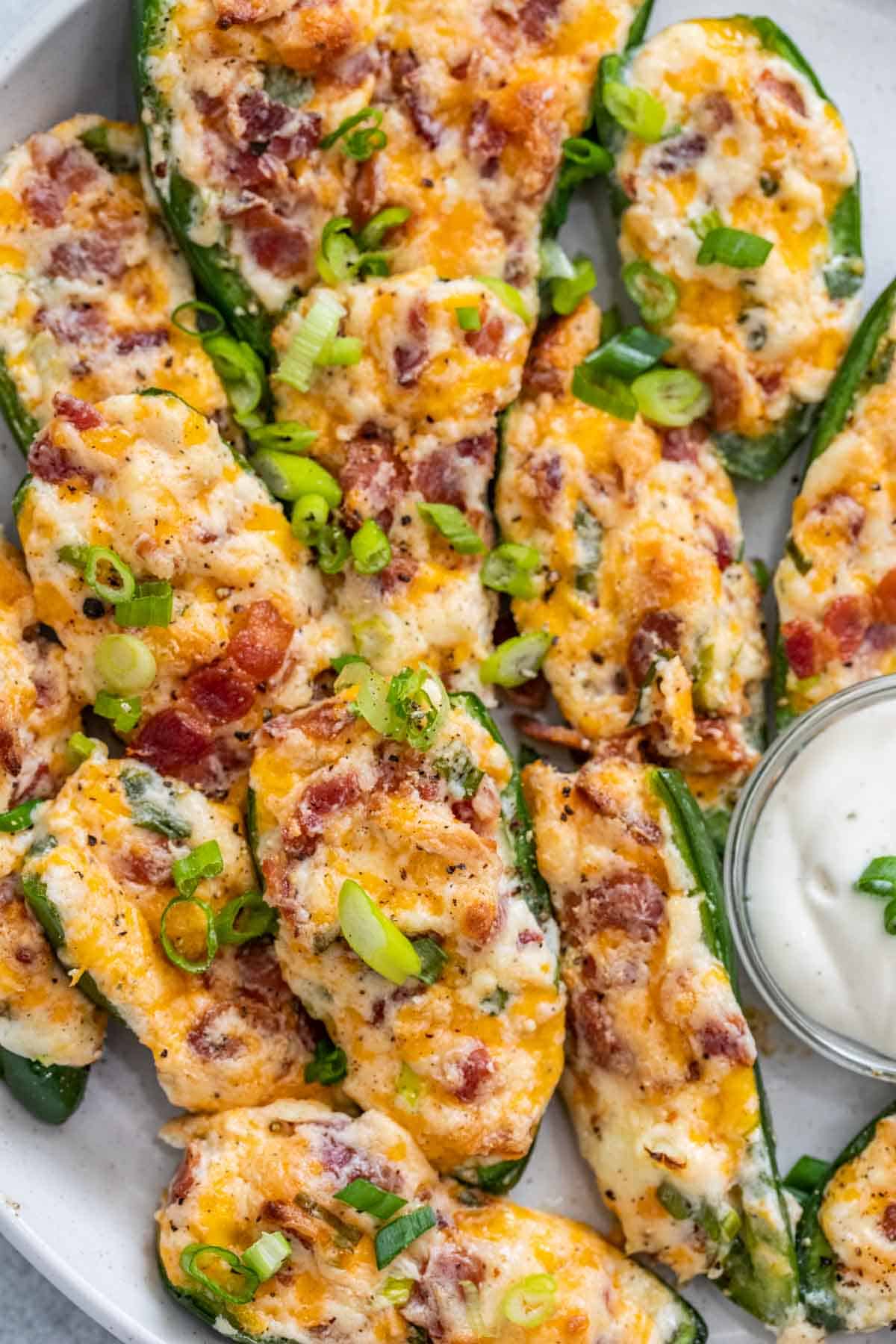 Stuffed jalapeno poppers laid out on a plate with a bowl of ranch topped with chopped green onion.