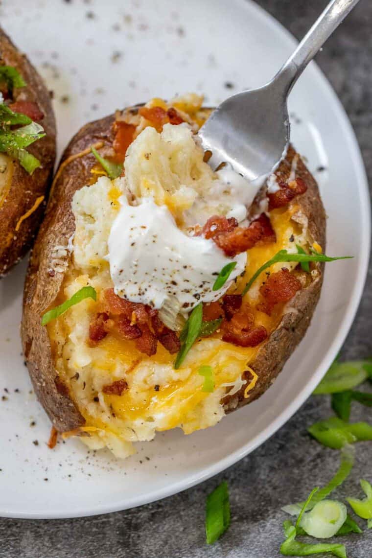 A twice baked potato on a white plate with a fork being dug into the potato. 