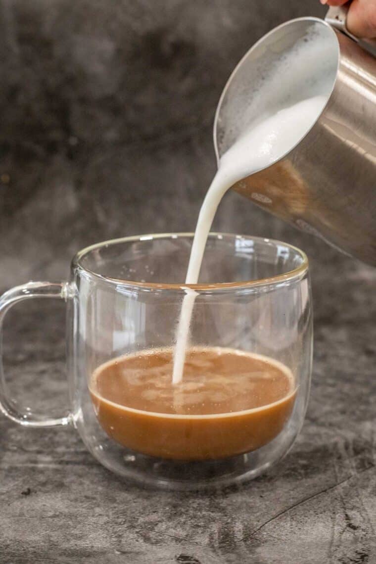 A glass mug of coffee with frothed milk being poured into it. 