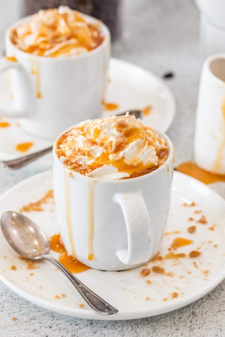 A white cup of caramel brulee latte topped with caramel drizzle and crushed candy. 