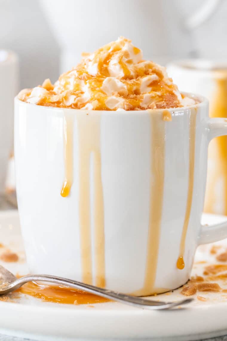 Caramel brulee latte in white mugs on white plated topped with caramel drizzle and topped with candy. 