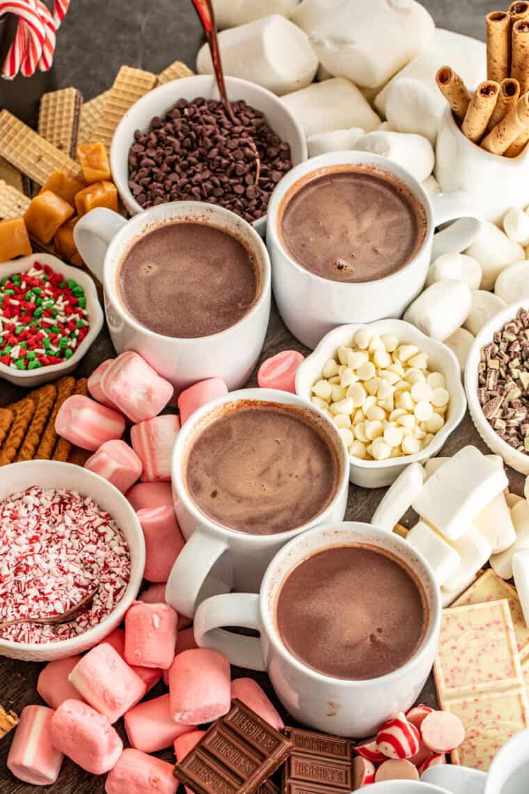 Four white mugs loaded with crockpot hot chocolate next to all the ultimate chocolate bar toppings. 
