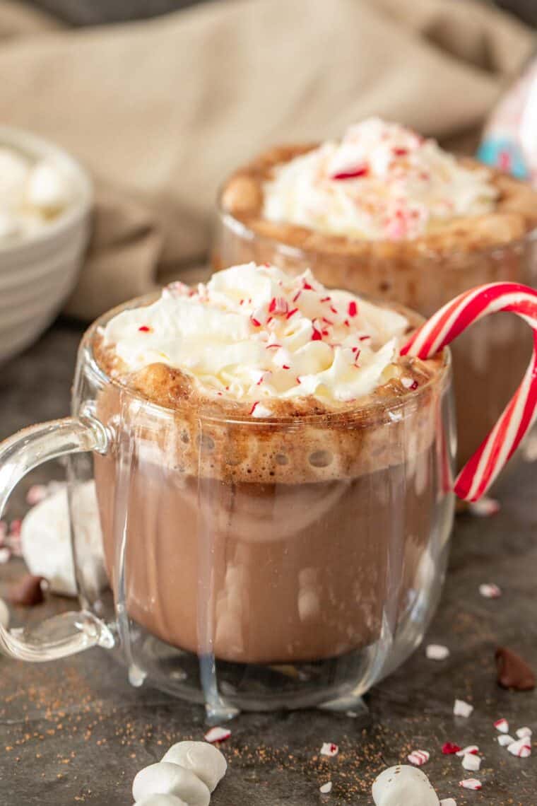 Peppermint hot chocolate in a glass cup topped with whipped cream and crushed peppermint with a candy cane in the cup. 