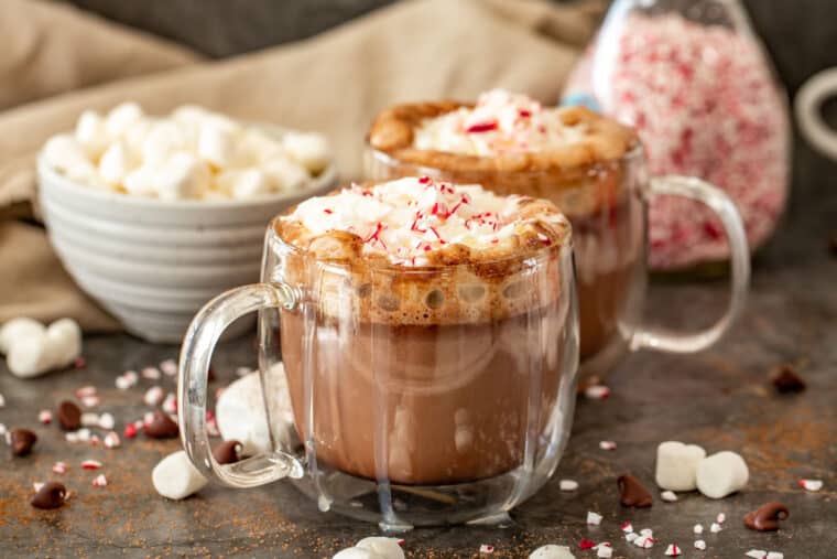 Hot chocolate in glass mugs topped with whipped cream and crushed peppermint next to a bowl of marshmallows. 