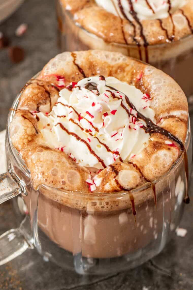 A glass cup of peppermint hot chocolate topped with whipped cream, crushed peppermint, and chocolate drizzle. 