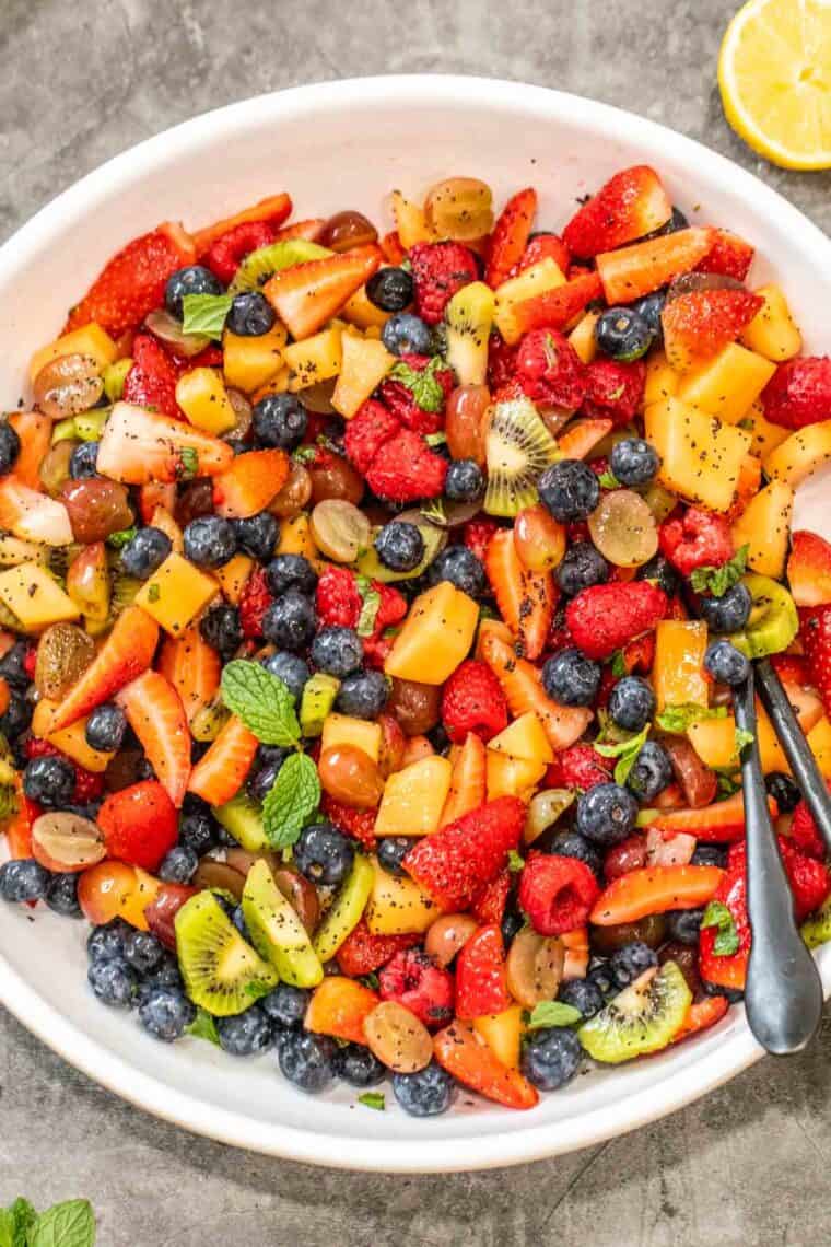 Fresh fruit salad in a white bowl with a metal spoon topped with fresh mint and poppy seeds.