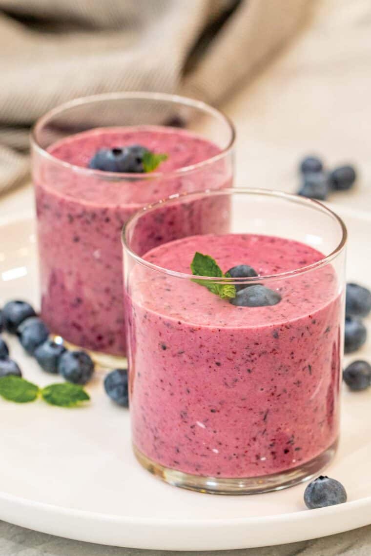 Blueberry smoothies in cups topped with fresh blueberries and fresh mint on a white plate.