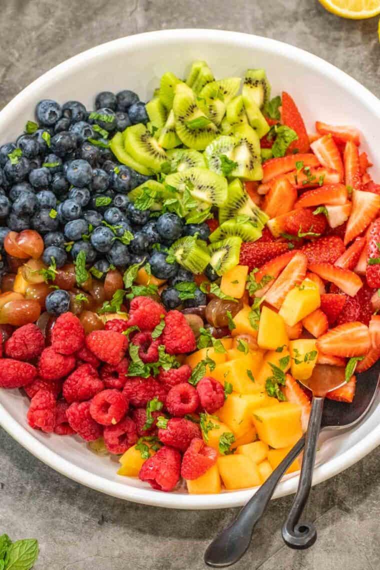 All the fruits needed for the best fruit salad laid out in a white bowl topped with fresh chopped mint. 