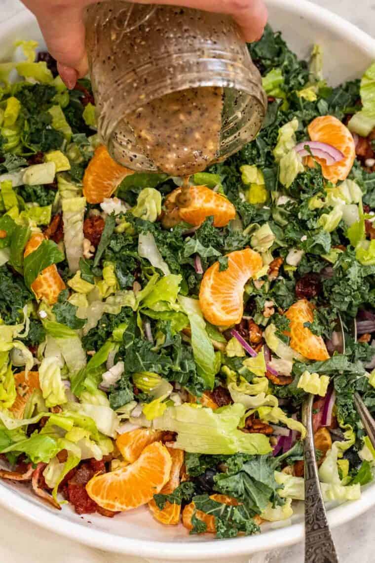 A white salad bowl with kale salad and the poppy seed dressing being poured into the salad. 