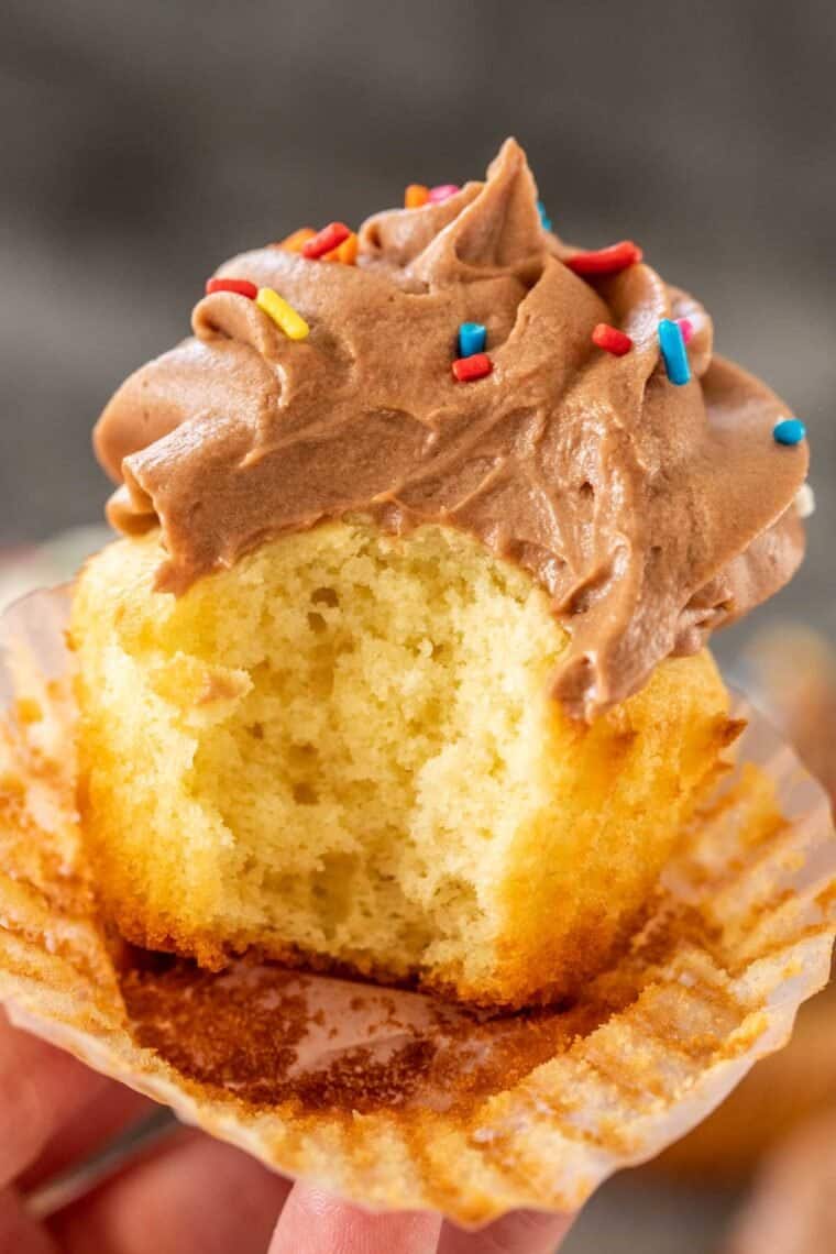 Vanilla cupcake with chocolate frosting with a bite being held by a person. 