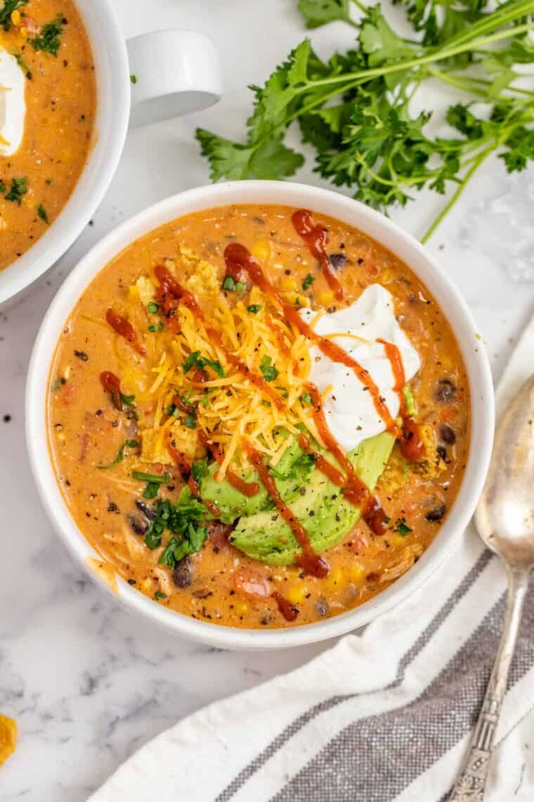 A white bowl with enchilada soup topped with shredded cheese, sour cream, avocado and drizzled with sriracha. 